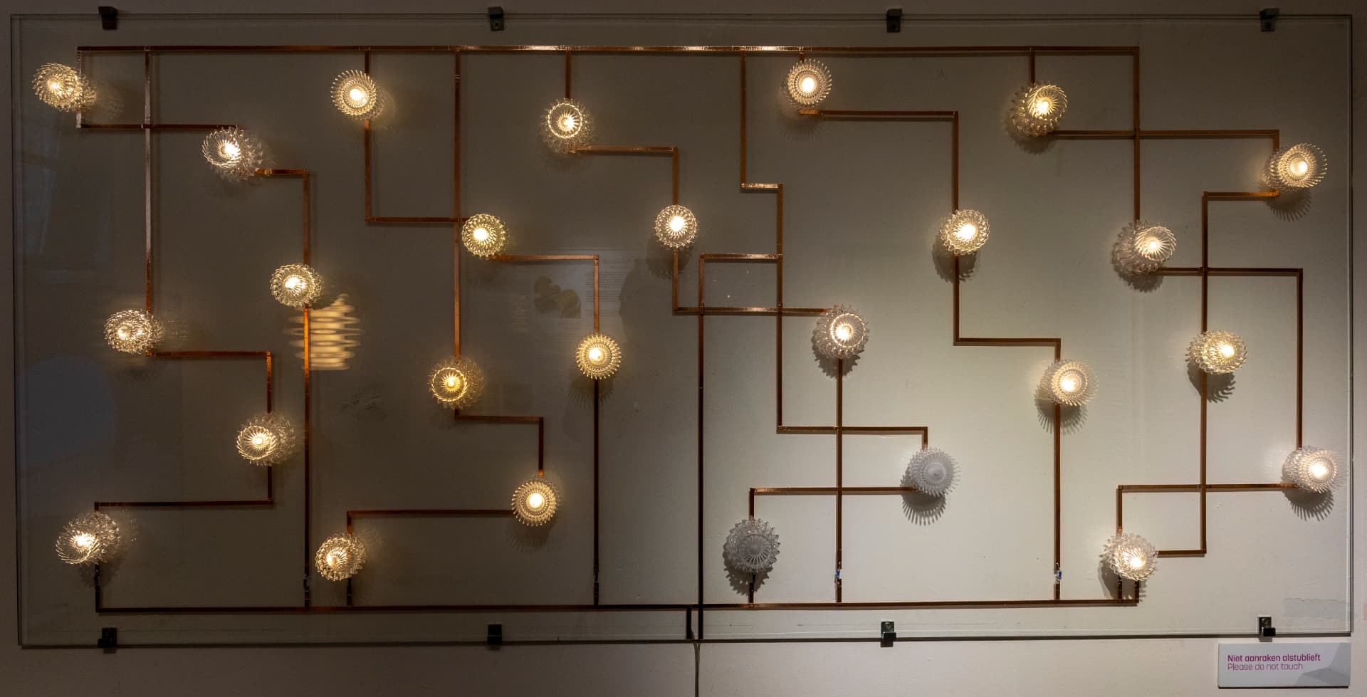 Wandinstallatie van losse verlichte glazen elementen, verbonden door koperen lijnen tot één zichtbaar patroon.